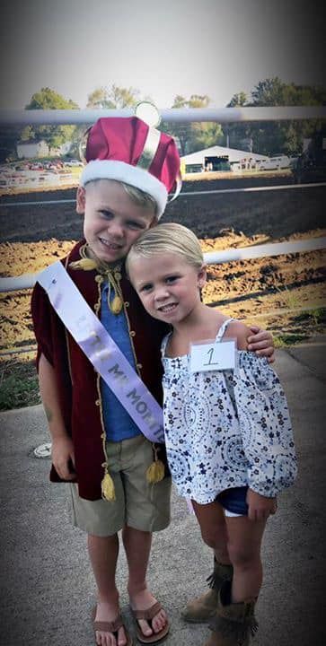 Two children posing outdoors, one wearing a red crown and sash, with a horse track in the background