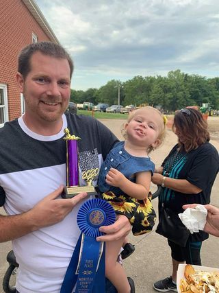 Man holding trophy and blue ribbon with smiling child outdoors at a sports event