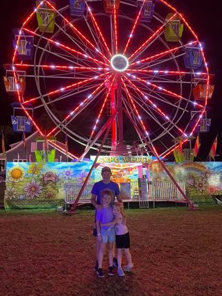 Two children posing in front of a brightly lit Ferris wheel at a nighttime fair.
