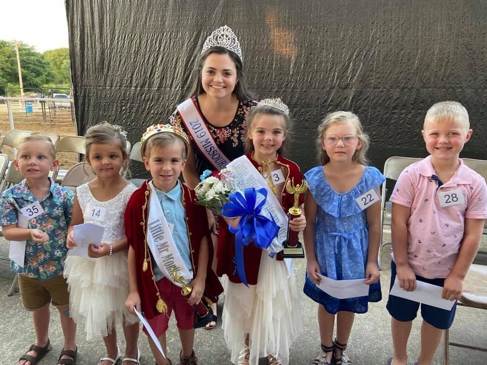 Woman with crown and sash holding bouquets, standing behind a row of children in sashes and pageant outfits