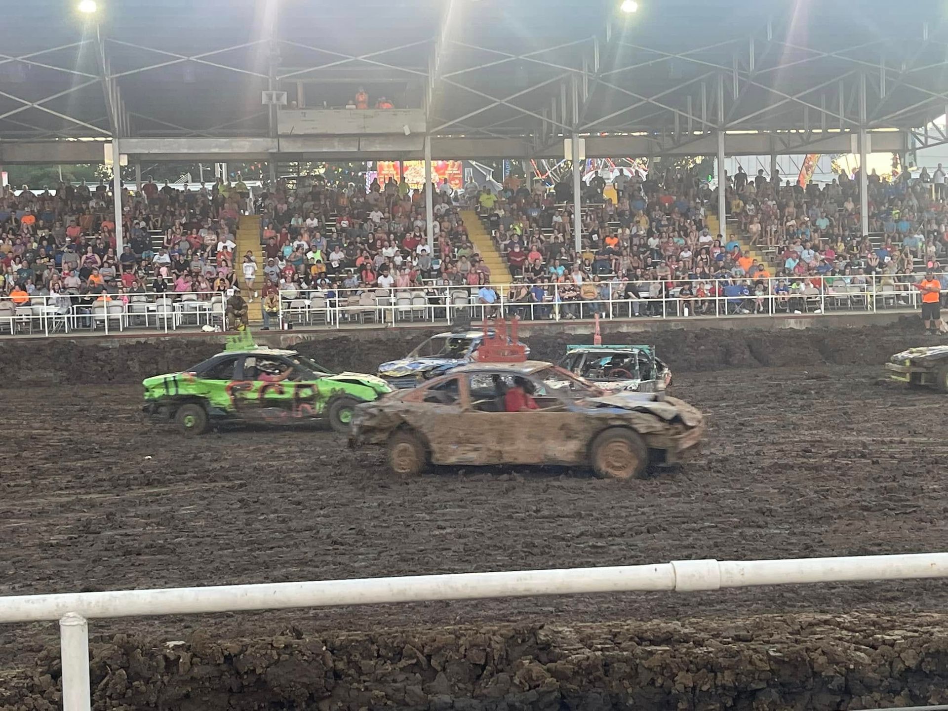 Mud-covered demolition derby cars racing in an indoor arena with a crowded grandstand.