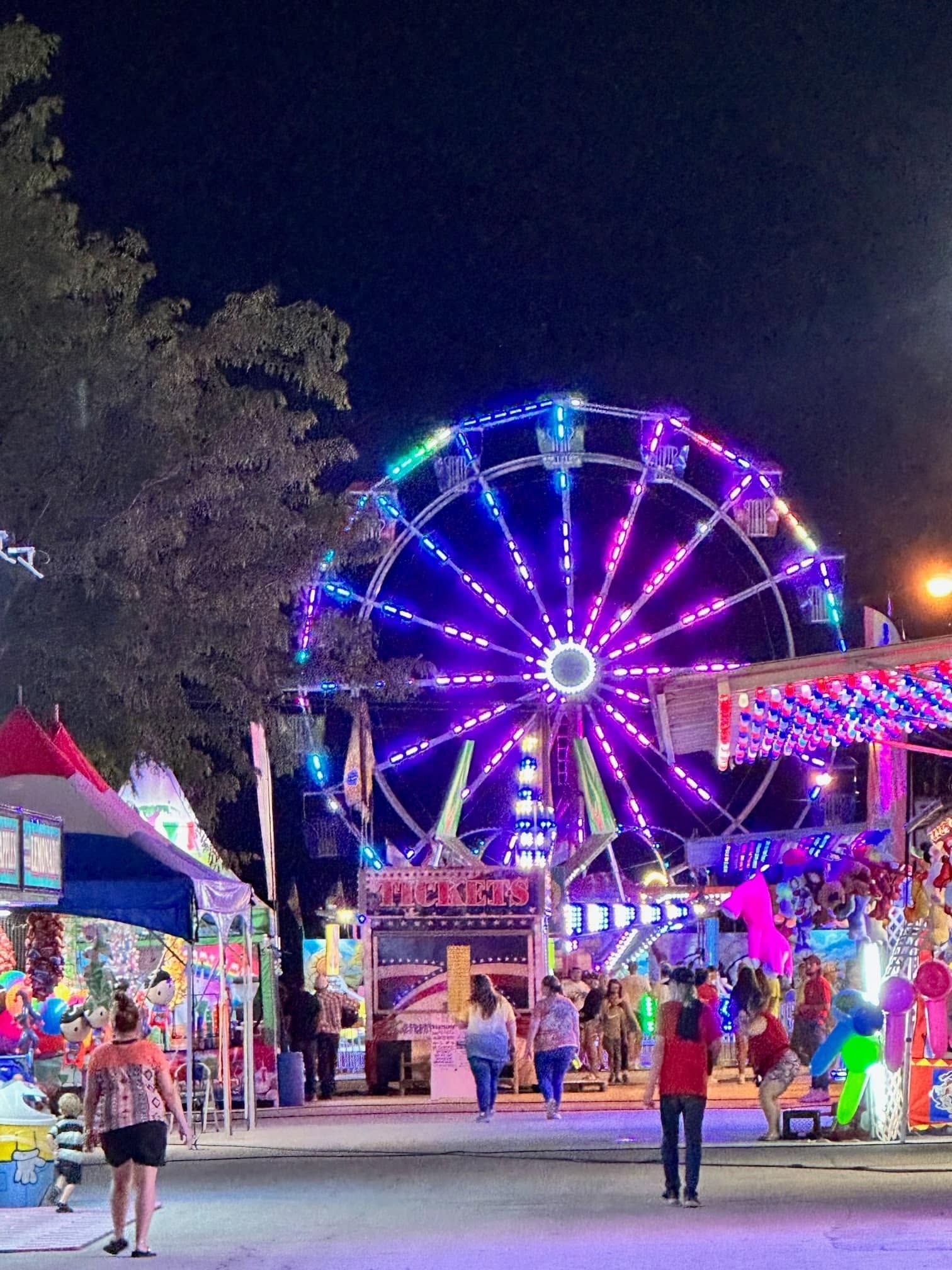 Night carnival walkway with a colorful Ferris wheel and bright vendor booths lit up at dusk