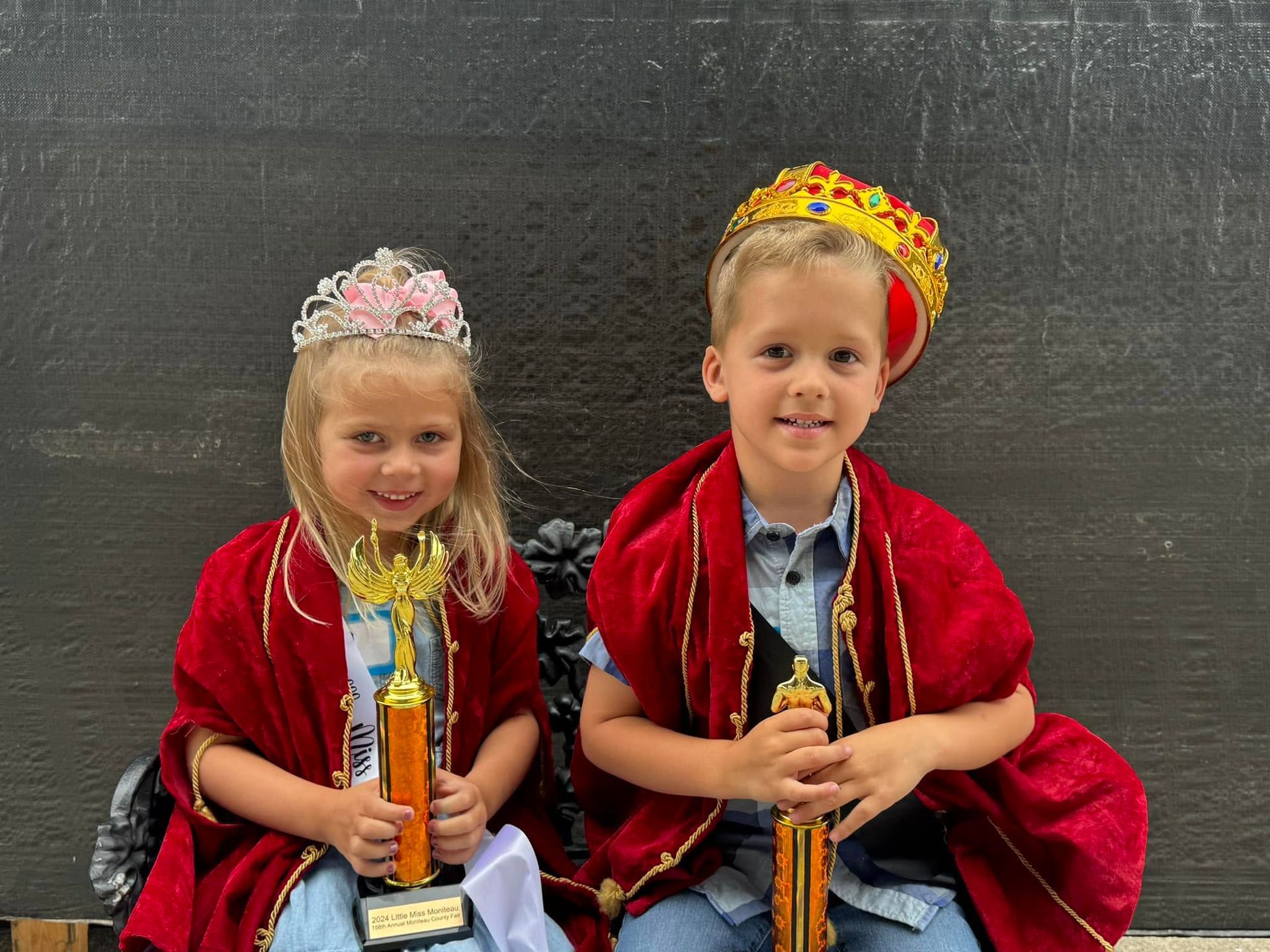 Two children in red costumes and crowns sit holding trophies against a dark backdrop.