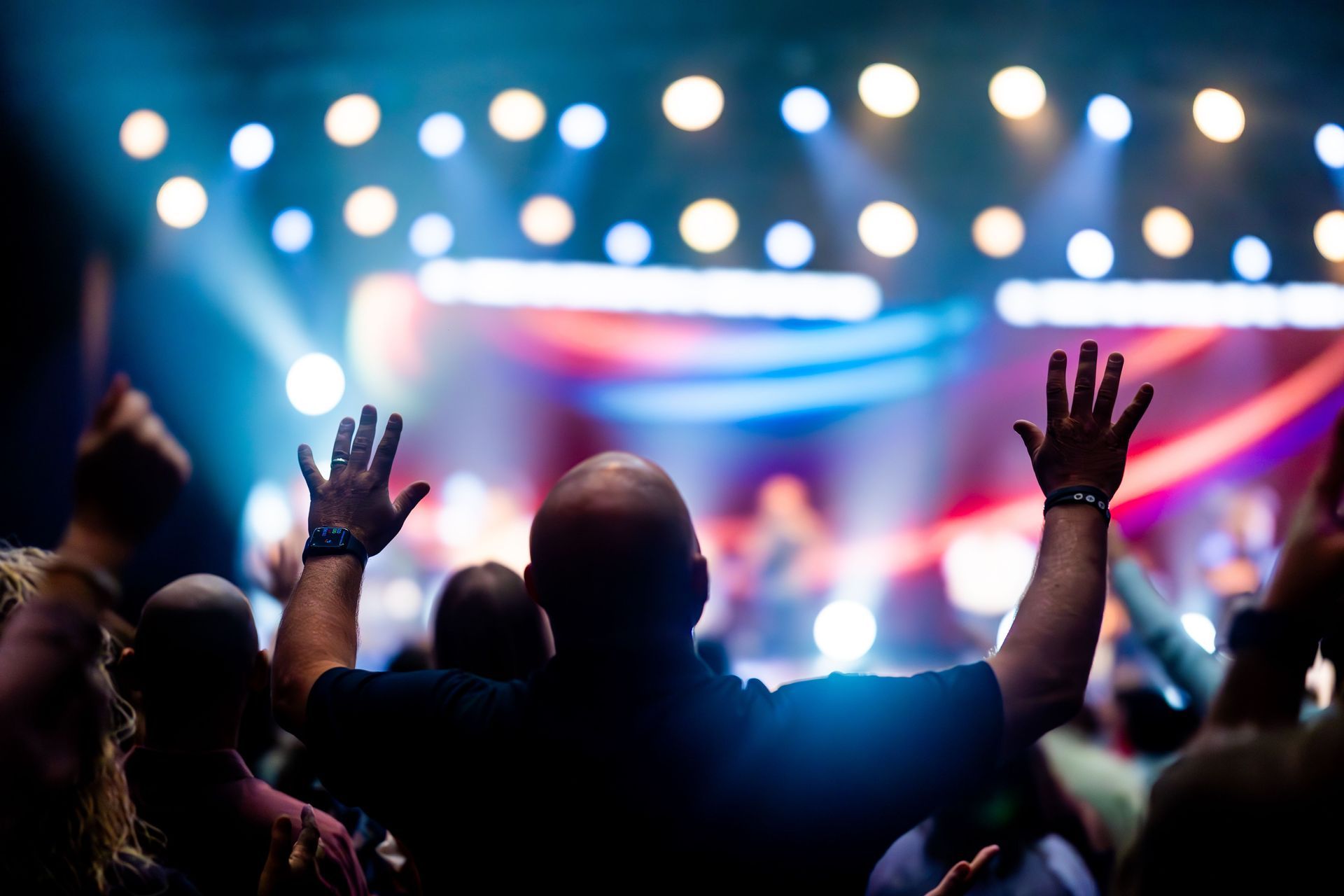 Crowd with raised hands at a concert, bright stage lights and colorful bokeh in the background