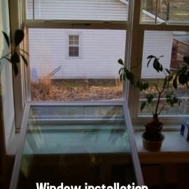 A large glass window box shelf installed into a house window, featuring a potted plant and view of a neighboring home.