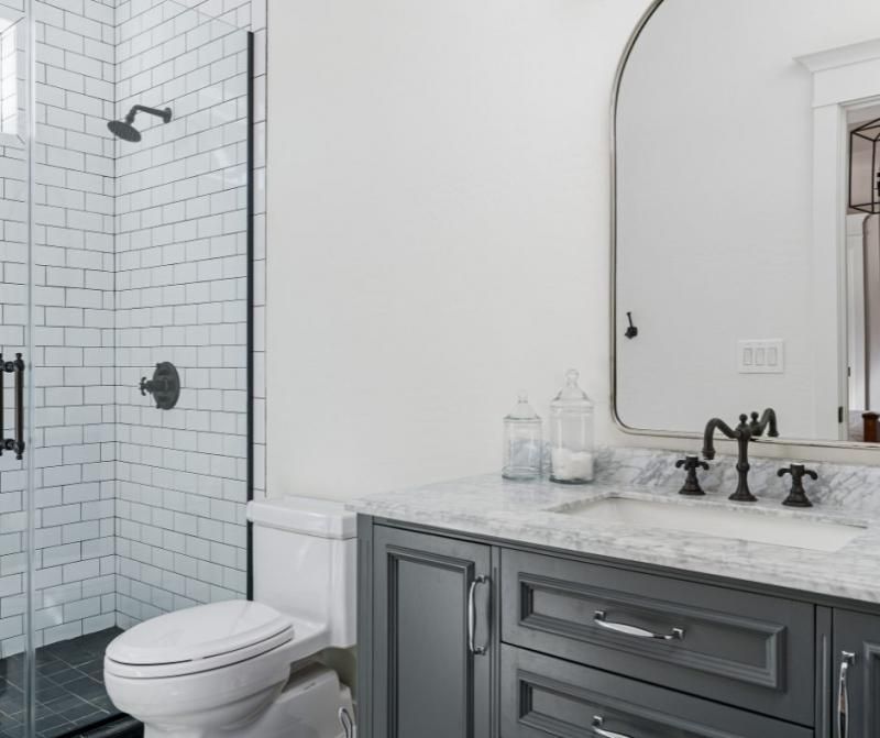 A modern bathroom featuring a charcoal gray vanity, white subway tile shower, and an arched mirror with black accents.
