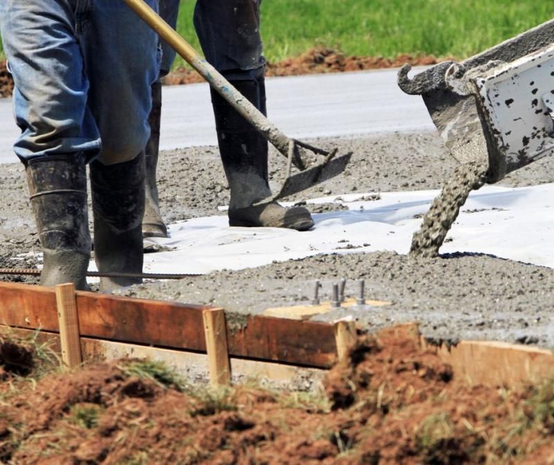 Construction workers pour and level fresh concrete onto a prepared site framed with wooden boards.