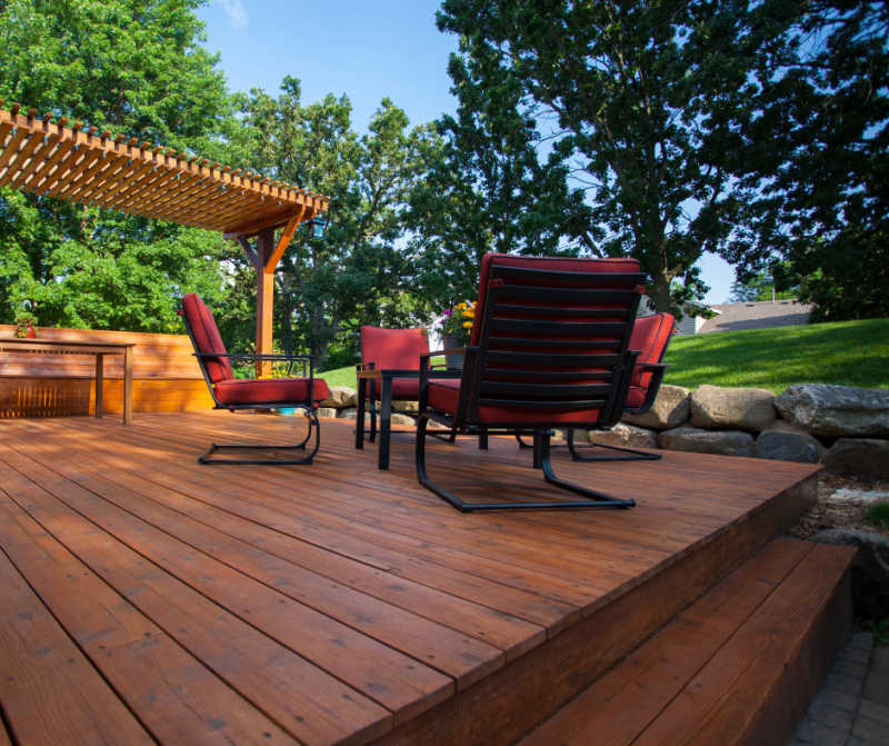 A wooden patio deck with matching chairs with red cushions sits under a pergola in a backyard with trees and a lawn.