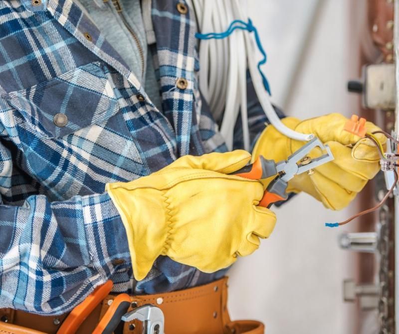 An electrician in a blue plaid shirt and yellow work gloves uses pliers to strip electrical wires near a wall outlet.