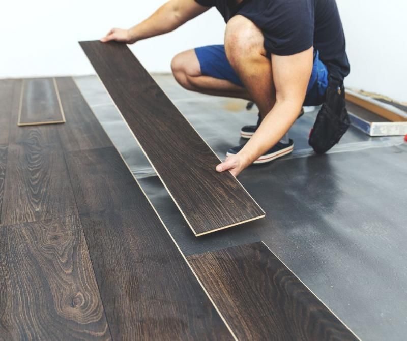 A person crouches while installing dark wood-look planks on a floor, connecting one piece to the existing layout.