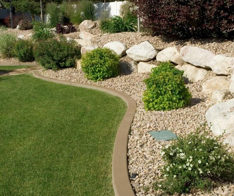 A manicured lawn bordered by a smooth concrete edge, adjacent to a rock garden with bushes and light-colored boulders.
