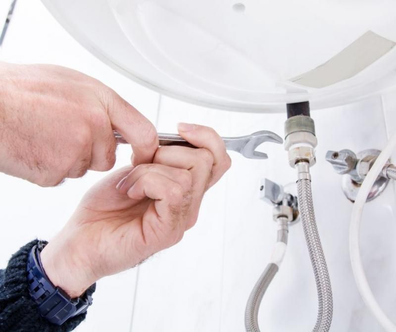 Hands using a metal wrench to tighten a fitting on the underside of a white appliance with braided water hoses.