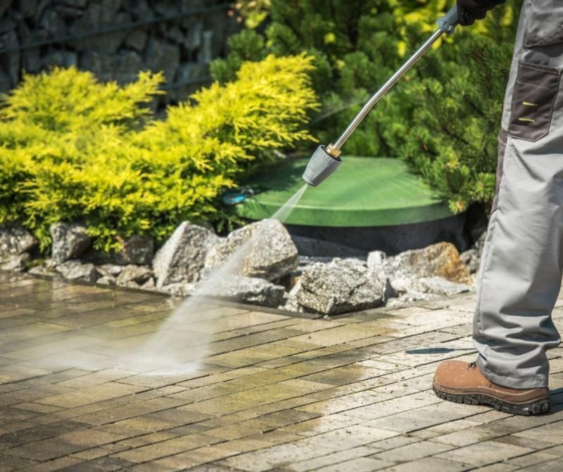 A person in gray work pants pressure washes a stone patio in a yard with green plants and rocks.