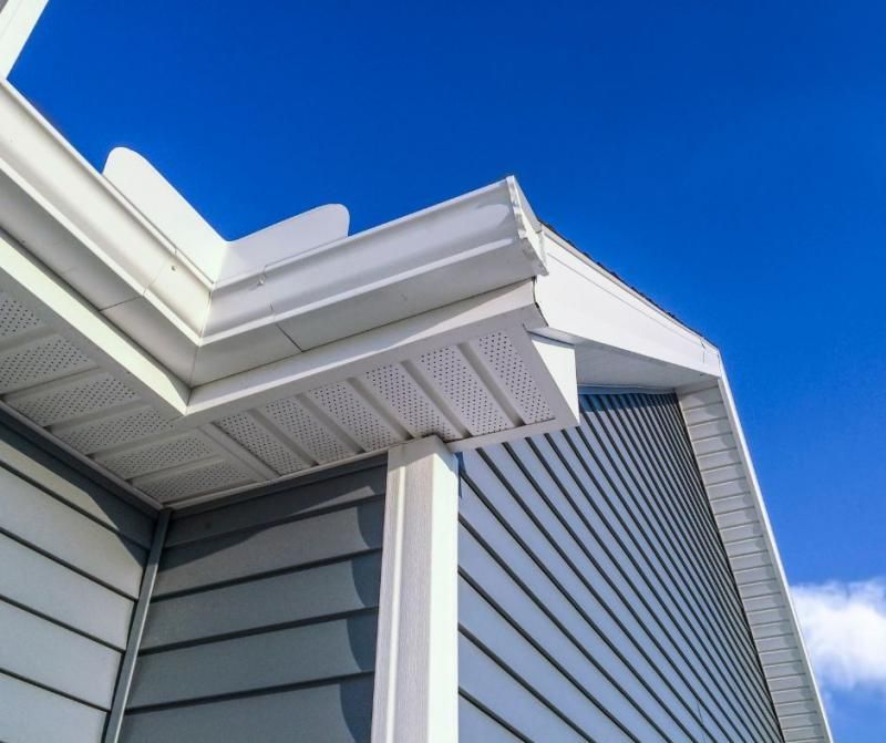 The corner of a building exterior showing light gray siding, white soffits, gutters, and a vertical trim against a blue sky.