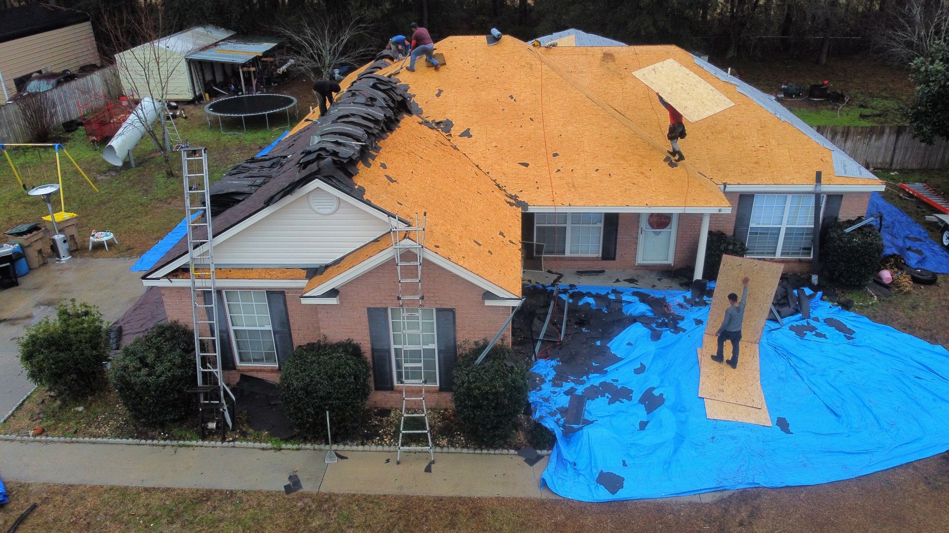 Roofers replacing a shingled roof on a brick home, covered in blue tarps.