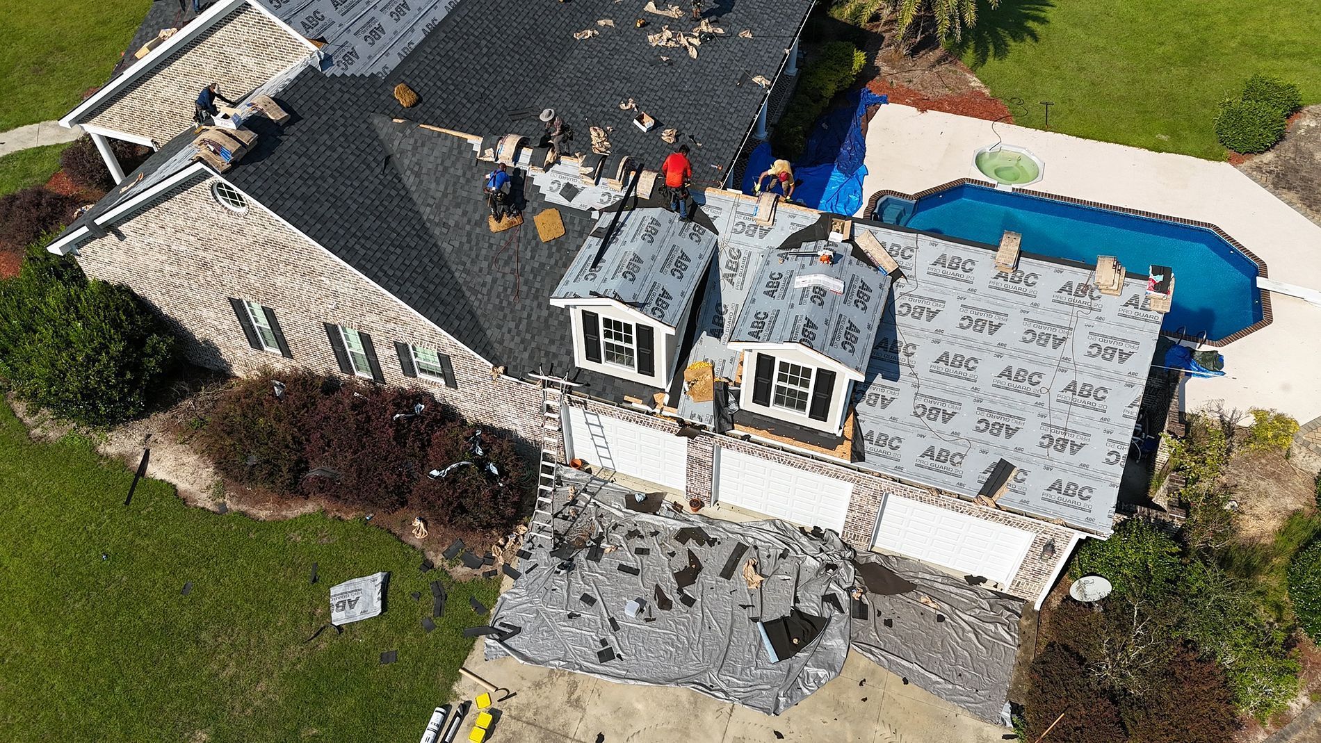 Aerial view of a house with roofers working on several roof sections, blue pool nearby.