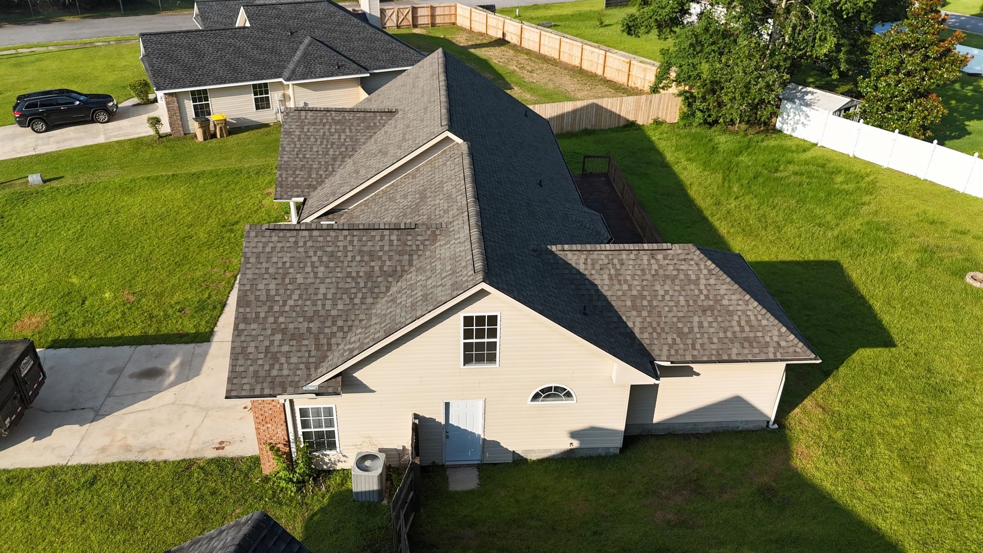 An aerial view of a house with a gray roof on a green lawn. A driveway and fence are visible.