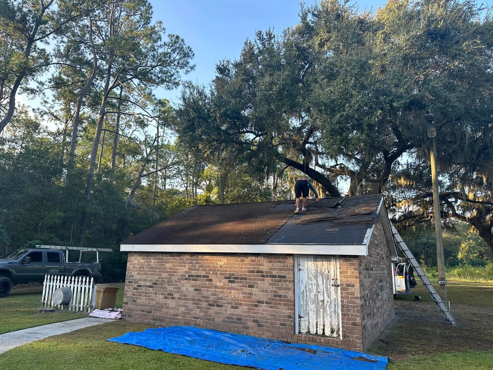 Person on a brick building roof, removing shingles. Blue tarp below. Trees in the background.