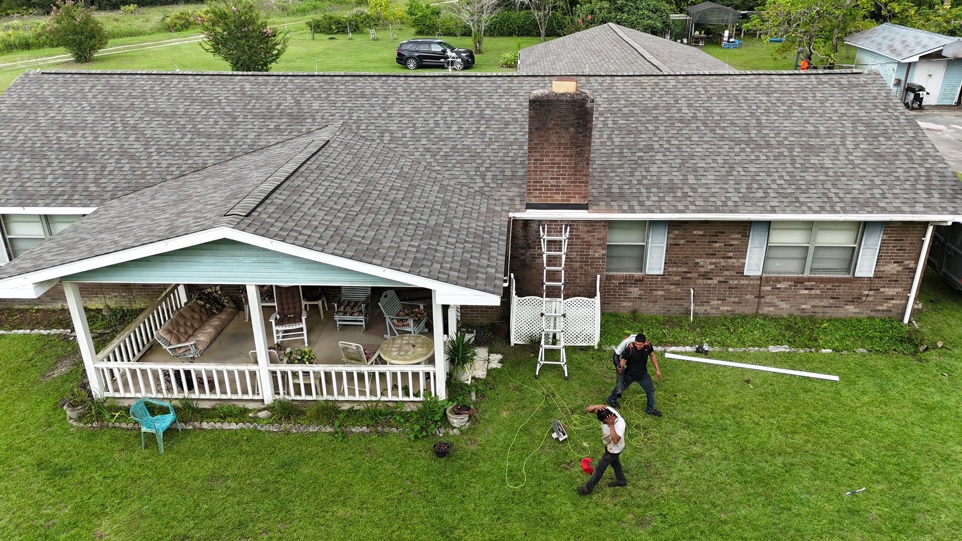 Overhead shot: Roofers working on a brick house with green lawn, ladder, and parked cars.
