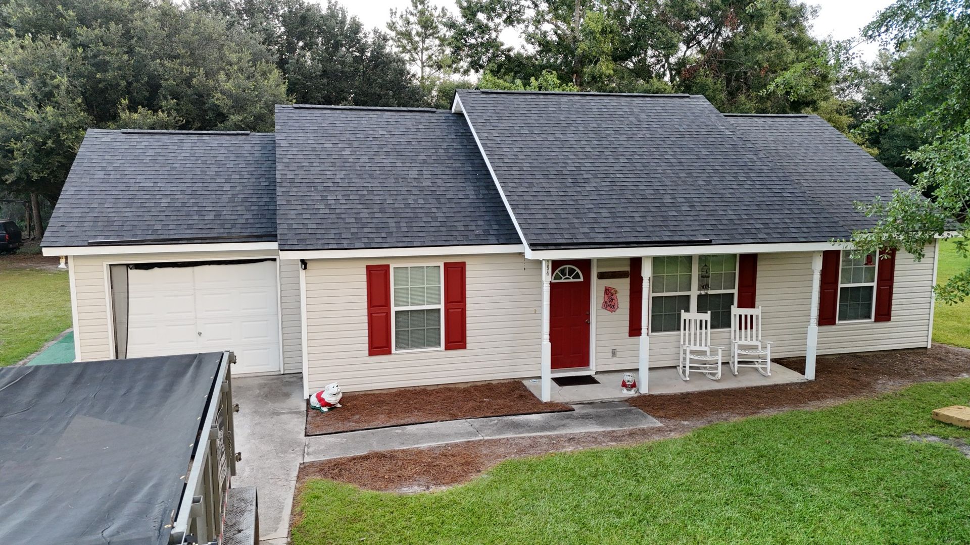 A light beige house with red shutters and a red door, and a gray roof. A black trailer is parked in front.