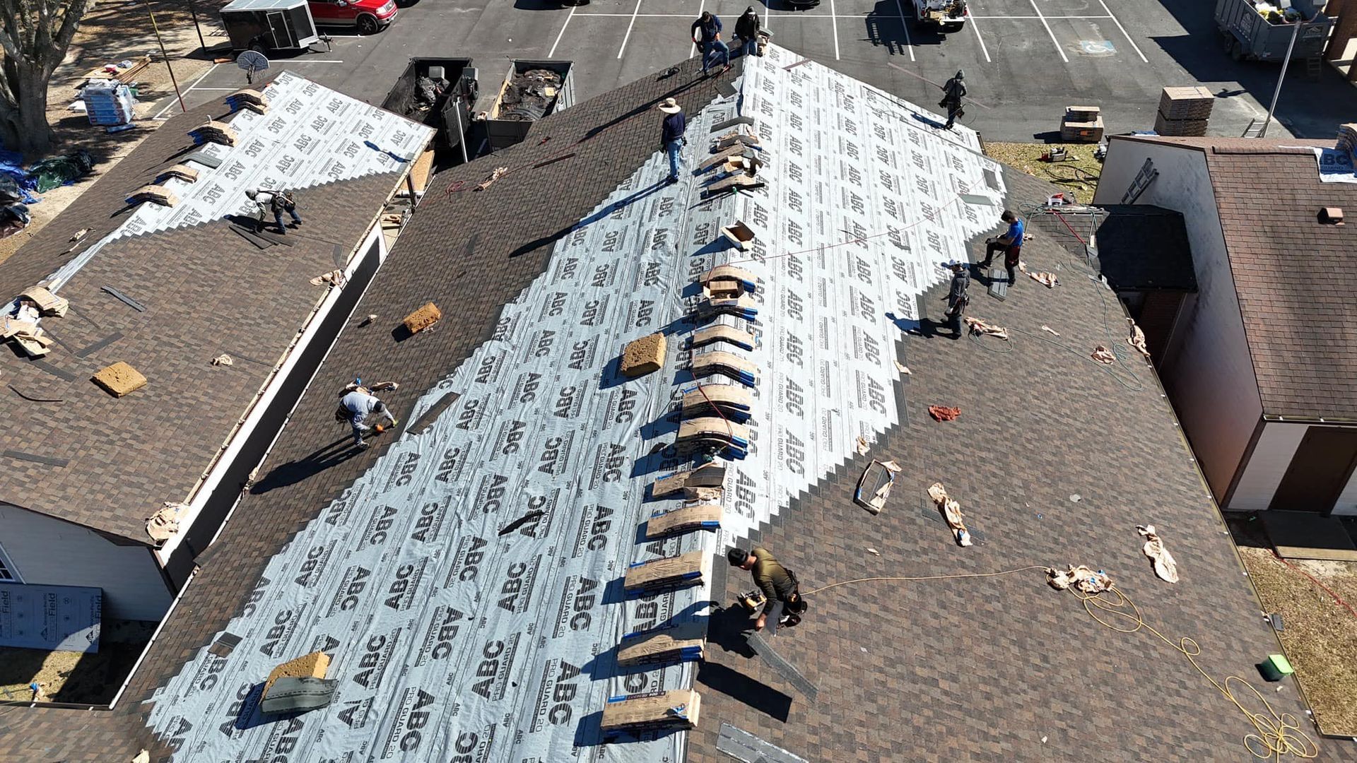 Roofers working on a brown shingled roof, installing protective underlayment on a sunny day.