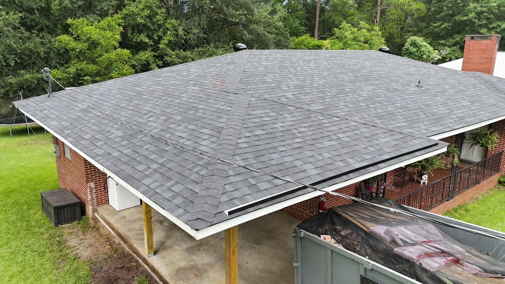 A dark grey shingled roof on a small building with a porch. Green trees in the background.