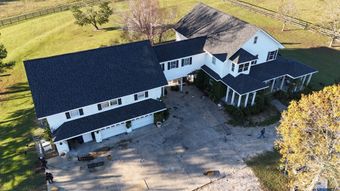 Large white house with black roof, a large concrete driveway, and surrounding green fields.