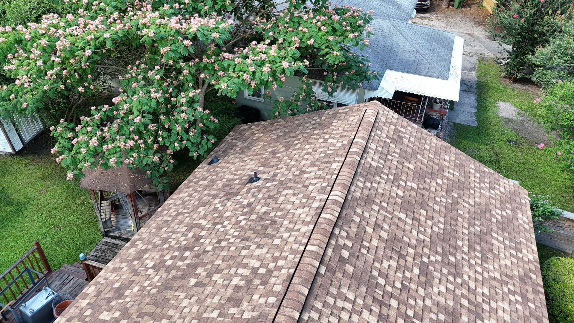 Aerial view of a brown shingled roof, surrounded by green grass and a flowering tree.