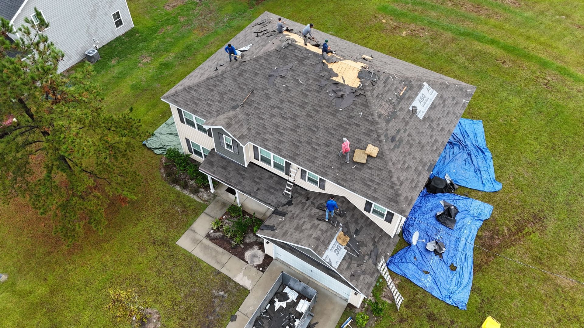 Aerial view of a two-story house with roof damage. Workers repair the roof while blue tarps protect the structure.