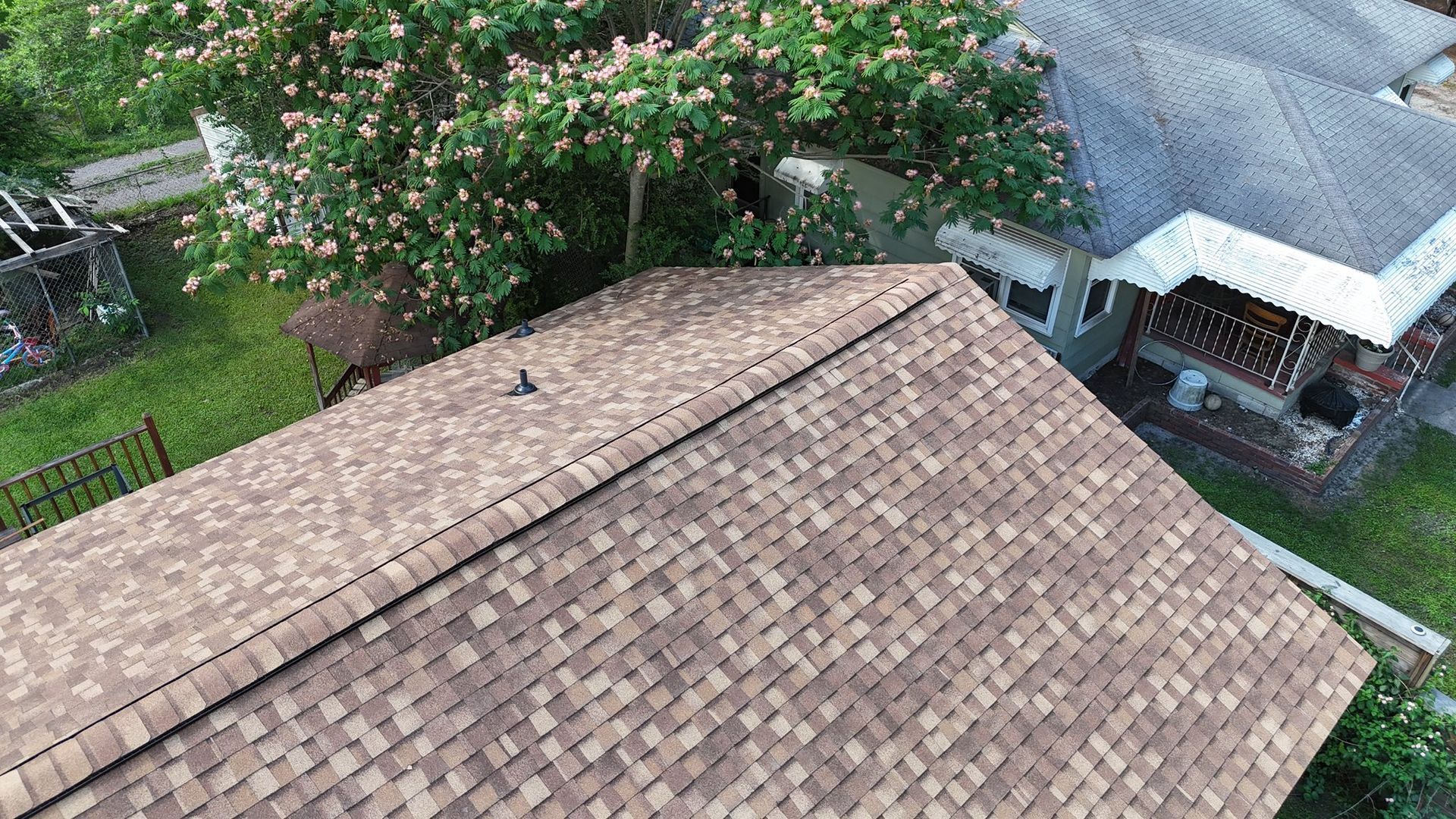 Aerial view of a brown shingle roof with a house next to trees and another building.
