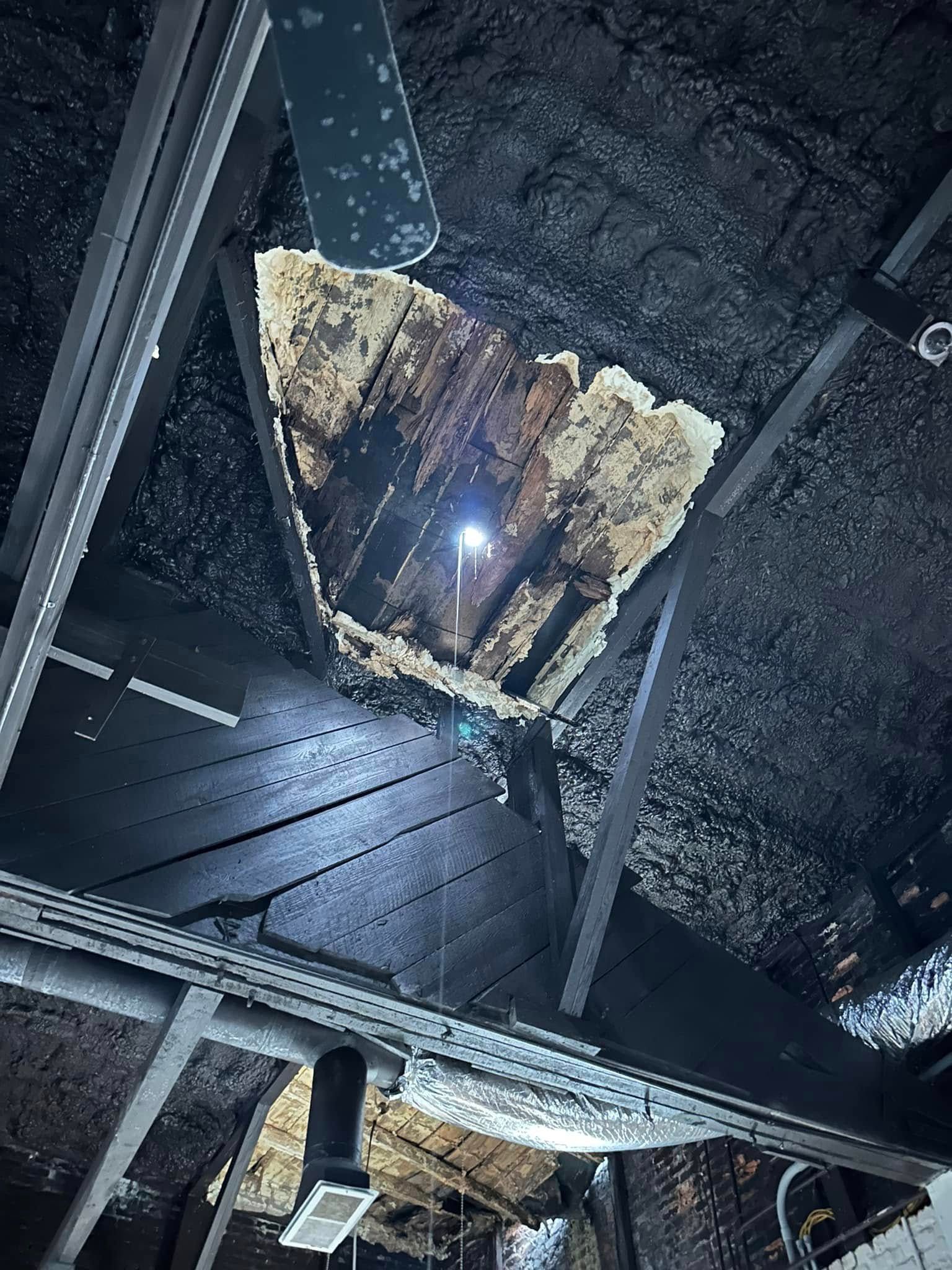 A hole in a dark ceiling, revealing light. Charred wood is visible around the opening.