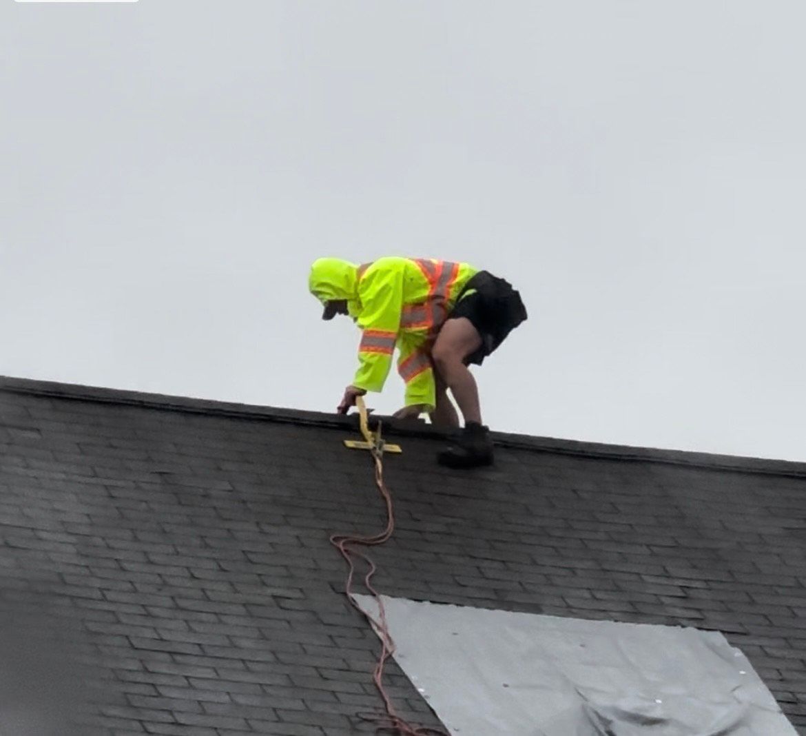 Worker in yellow safety gear on a sloped roof, connecting a safety harness. Cloudy sky.