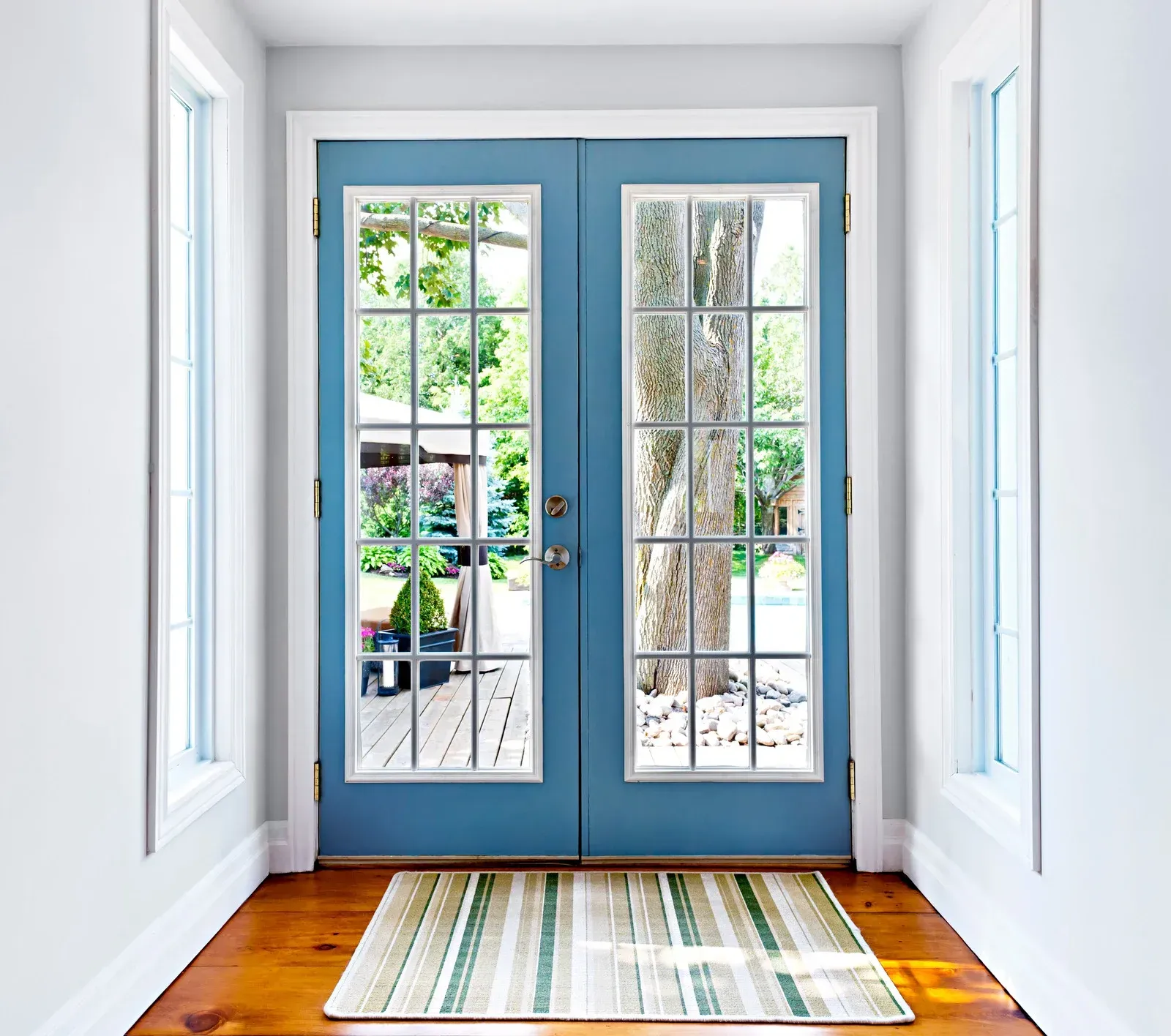 Blue French doors with glass panes, flanked by windows, opening to a backyard, and a rug on a wooden floor.
