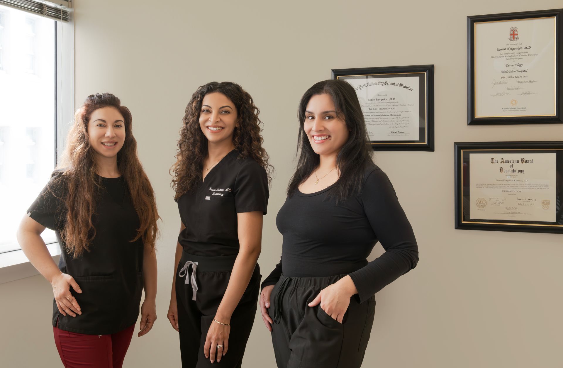 Three women standing in an office with diplomas on the wall. They wear black scrubs and tops. One has long wavy hair.