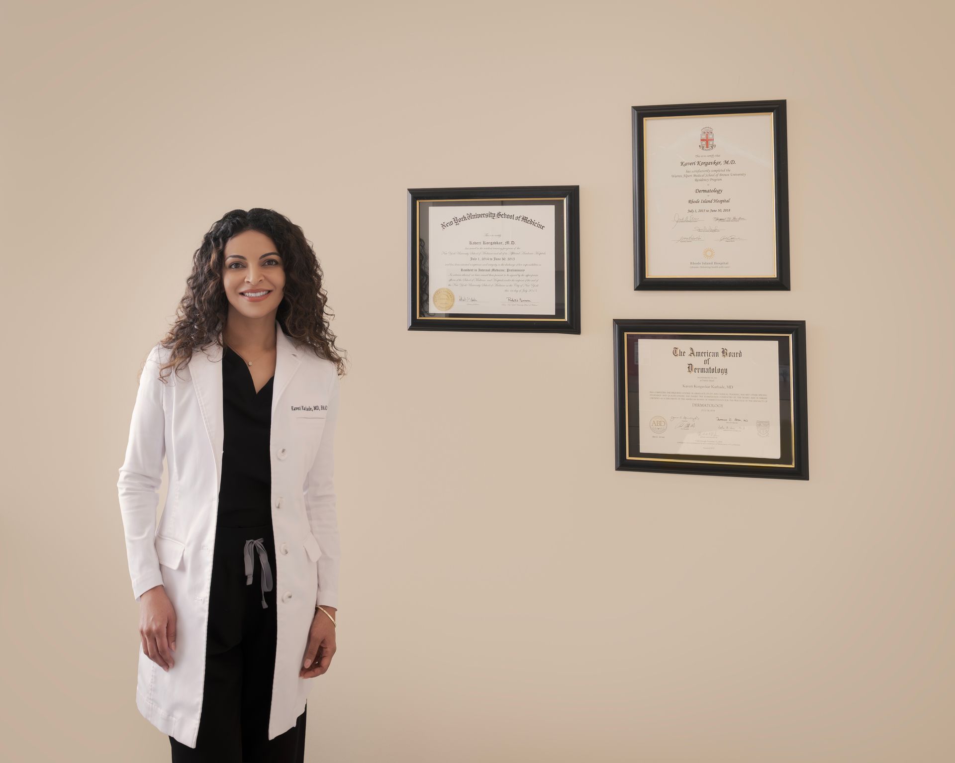 Woman in white coat smiles, standing next to framed certificates on a beige wall.