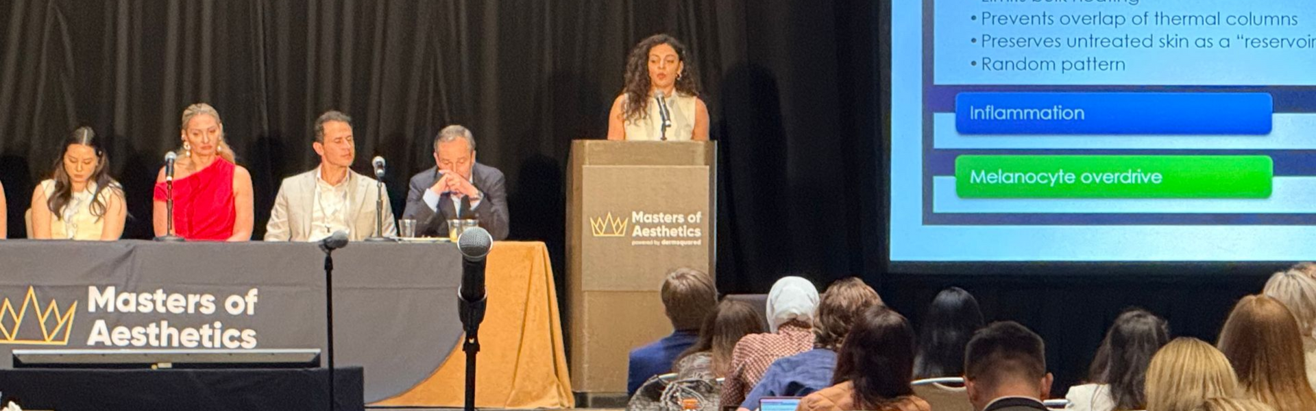 A presenter speaks from a podium at the Masters of Aesthetics event with a panel of four people seated at a table.