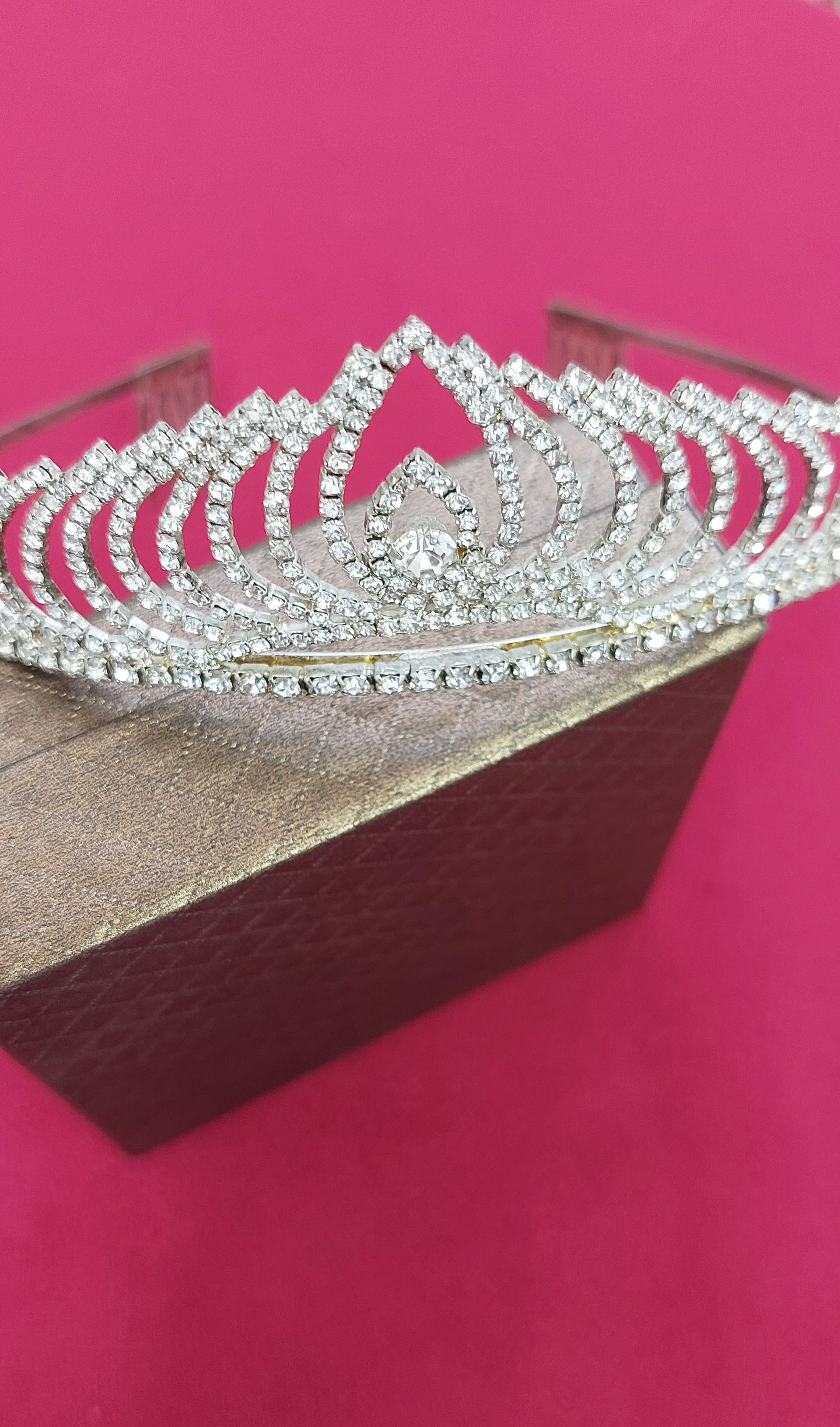 Tiara d'argento con strass scintillanti su una scatola dorata, su sfondo rosa.