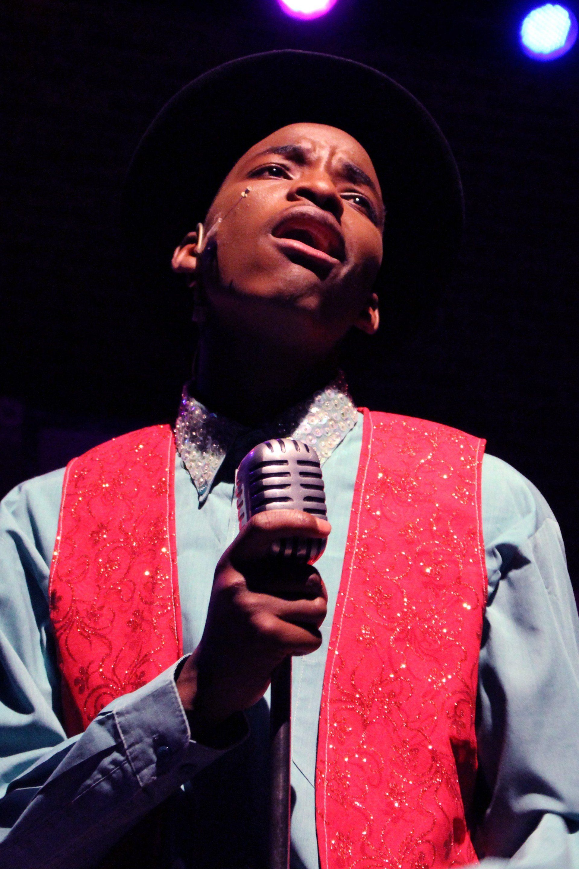 a boy singing into a microphone wearing a hat and a red vest
