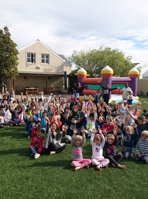 a group of children are sitting on the grass in front of a bouncy house .