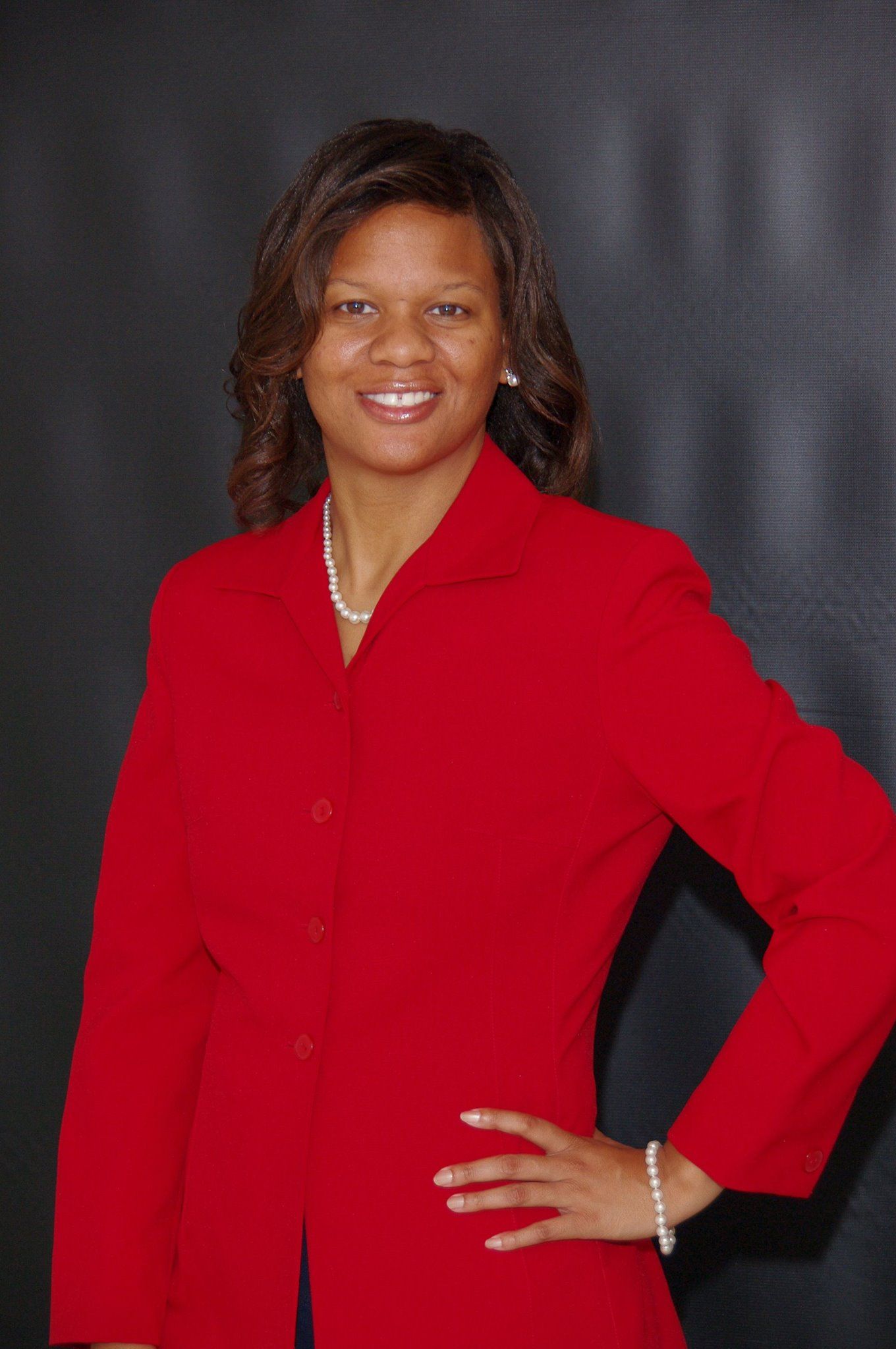 A woman in a red jacket and pearls is standing with her hands on her hips.