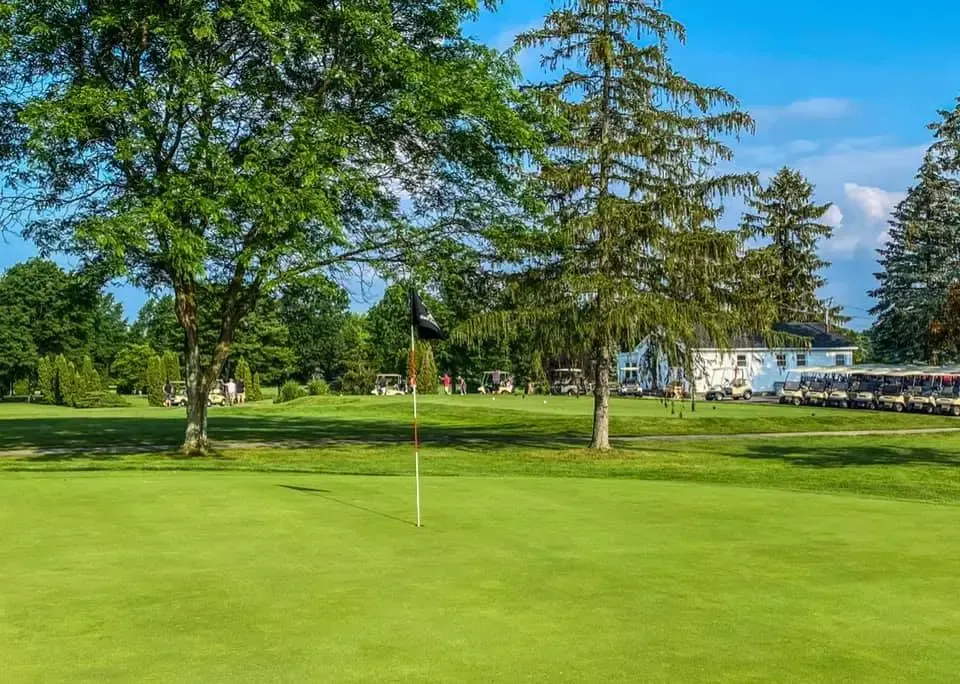 Green golf course with flag, trees, and white building in background under blue sky.
