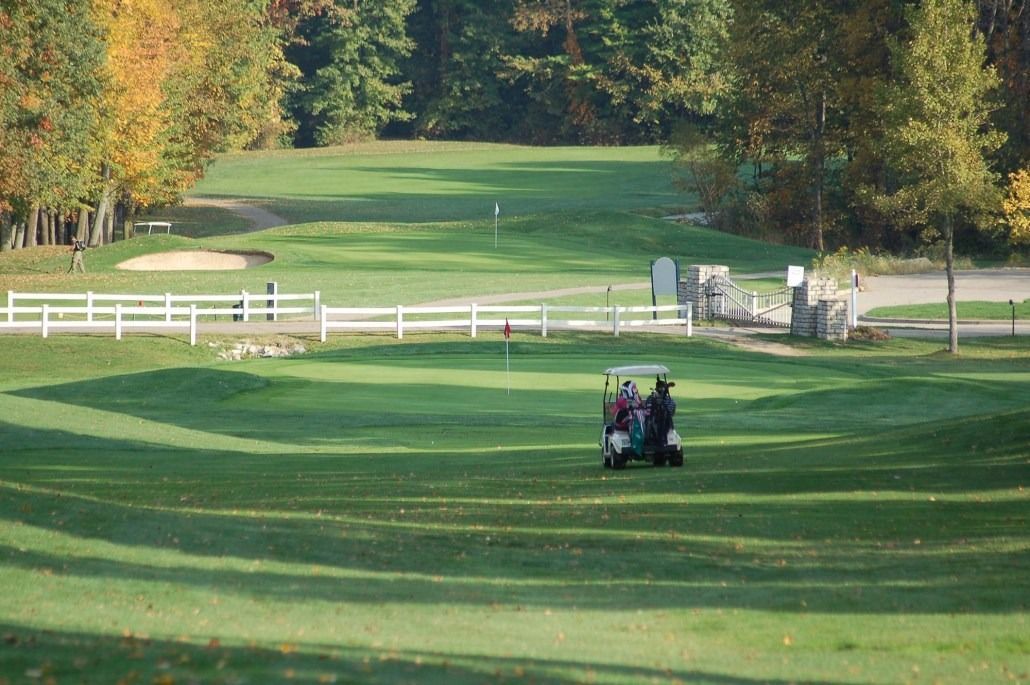 Golf course with a cart carrying two people on the green, fall trees in background.