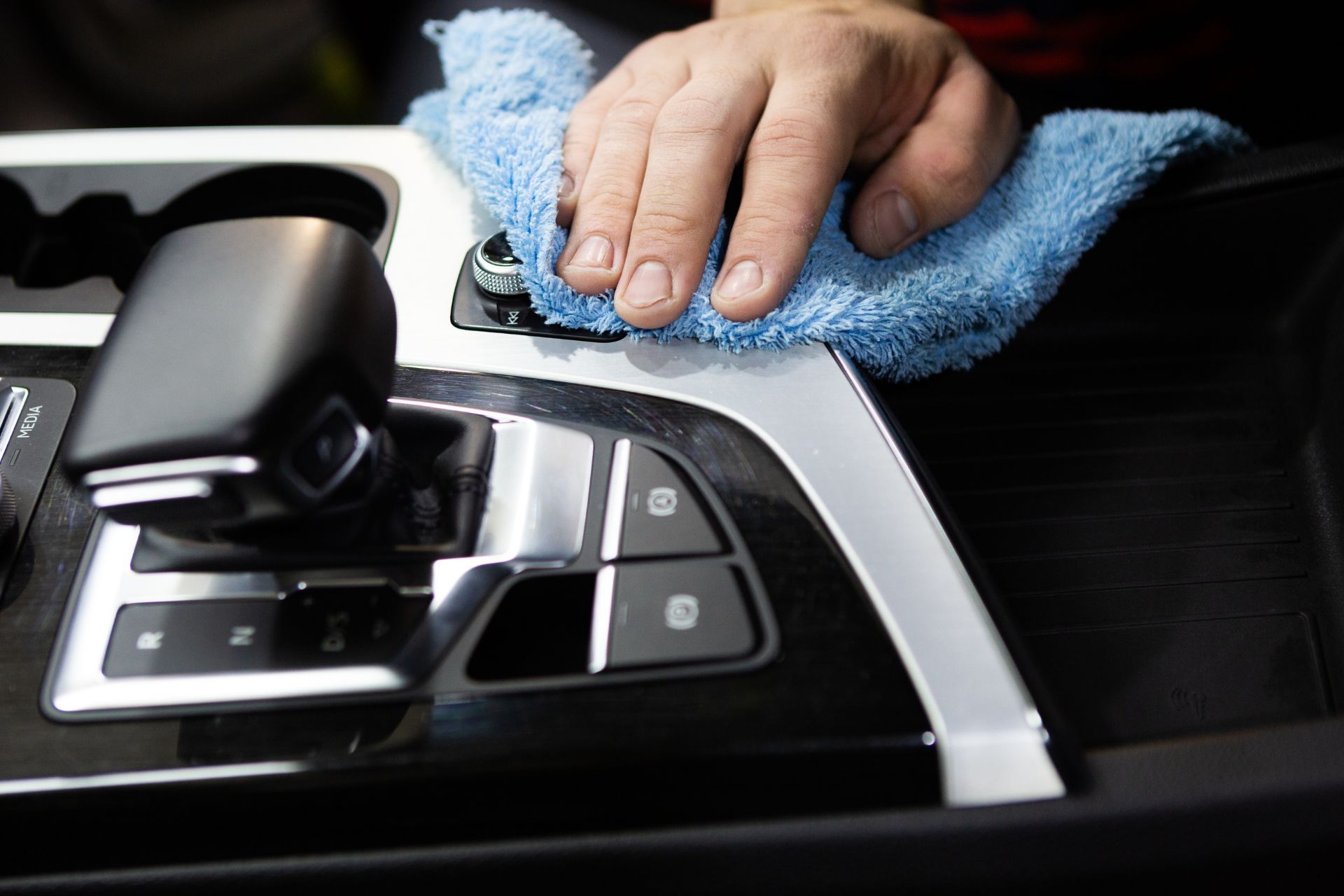 Close-up of a person's hand using a soft blue microfiber cloth to clean the glossy black center console of a car in Duluth, GA. The console includes a modern automatic gear shifter and various control buttons with metallic trim, reflecting meticulous auto detailing that enhances the vehicle's interior appearance.