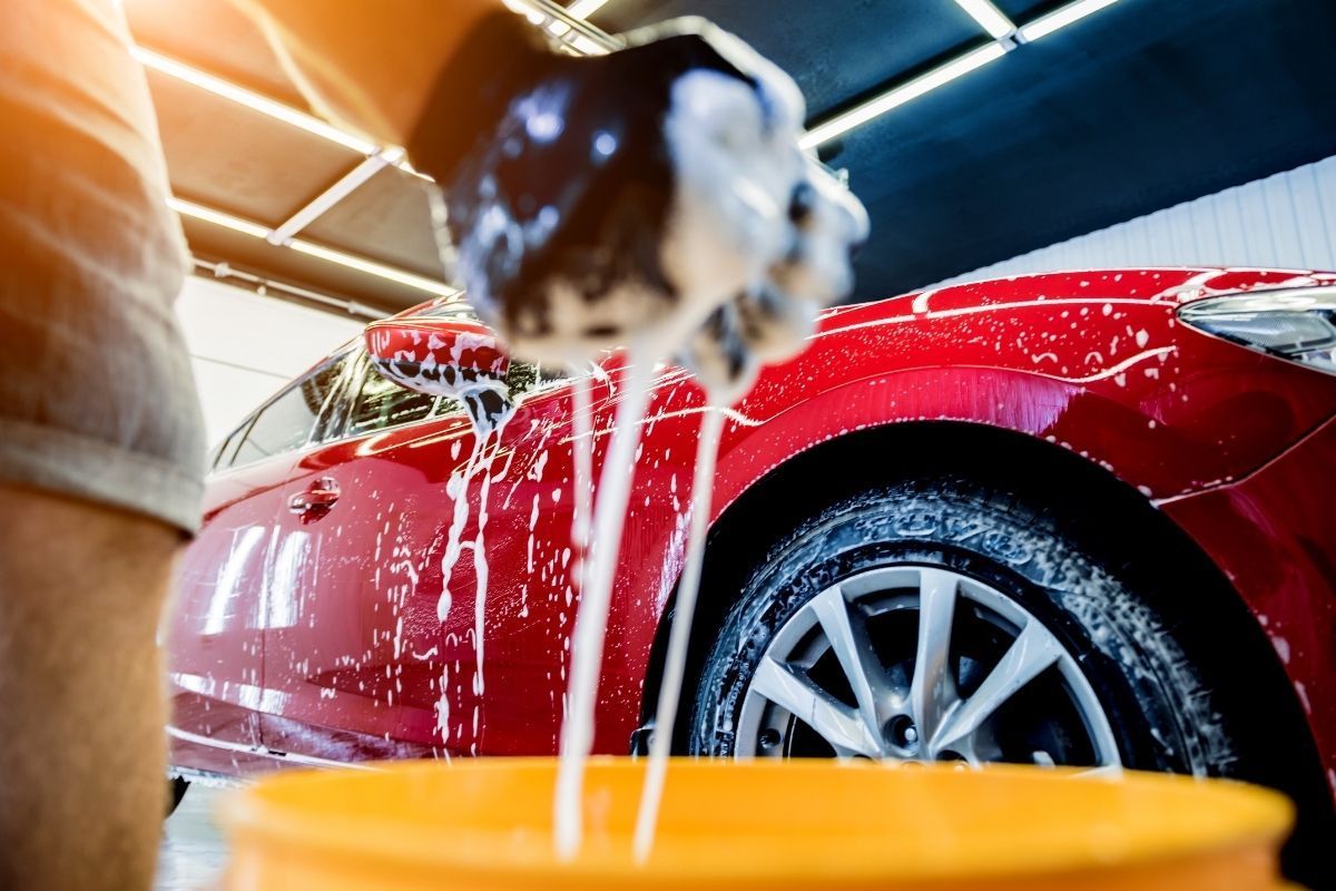 Close-up of a person washing a vibrant red car, with soapy water cascading over the vehicle's curved surface and wheel. The car is partly visible, emphasizing the sparkling clean bodywork and shiny alloy wheel. Suds drip onto the wet asphalt from a sudsy brush in the foreground, showing an active cleaning process. The indoor setting is lit by overhead fluorescent lights, suggesting a professional or home garage environment in Duluth, GA, dedicated to car maintenance.