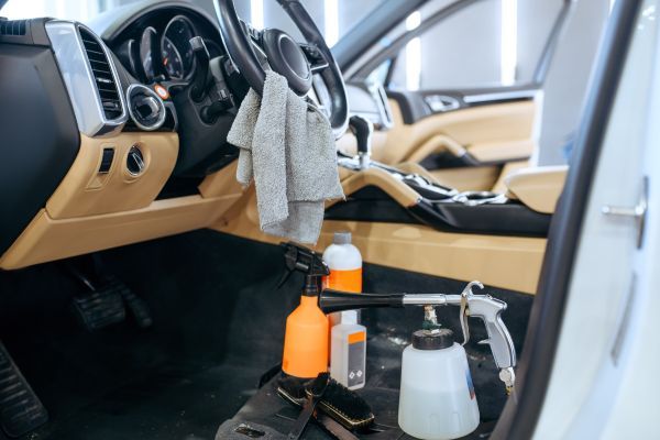 The image depicts the interior of a vehicle during a cleaning process, focusing on the driver's side with the door open. Inside, the car features a luxurious tan interior with leather seats. On the driver's seat, there is a gray microfiber cloth, suggesting an ongoing or just completed wipe down. In the foreground, an array of car detailing equipment is visible on the floor mat, including bottles of cleaning solutions, one with a bright orange sprayer, and various brushes with different bristle types designed for reaching into nooks and crannies. The presence of these tools indicates thorough interior detailing work. The car’s steering wheel, dashboard, and center console are visible, with clean surfaces reflecting meticulous care.
