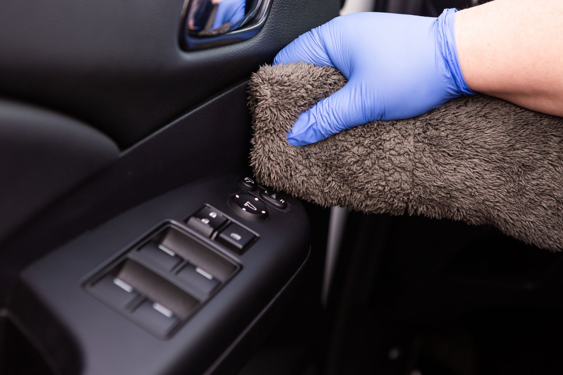 In a car in Duluth, GA, a person wearing blue gloves is diligently cleaning the black interior door panel with a fluffy gray microfiber cloth. The door has window and mirror adjustment controls, highlighting the thoroughness of the detailing service provided.