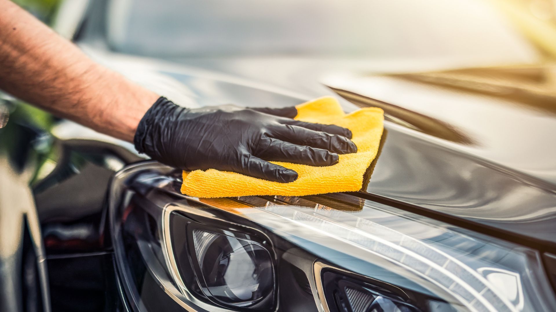 Close-up of a professional automotive detailer's hand, wearing a black glove, meticulously polishing a car's hood with a yellow microfiber cloth in Duluth, Georgia. The reflection on the shiny black paintwork indicates the effectiveness of the detailing work, exemplifying the care and attention to detail that goes into maintaining the vehicle's appearance. The image indicates the high-quality mobile detailing services available in Duluth, enhancing the aesthetic appeal and longevity of the car.