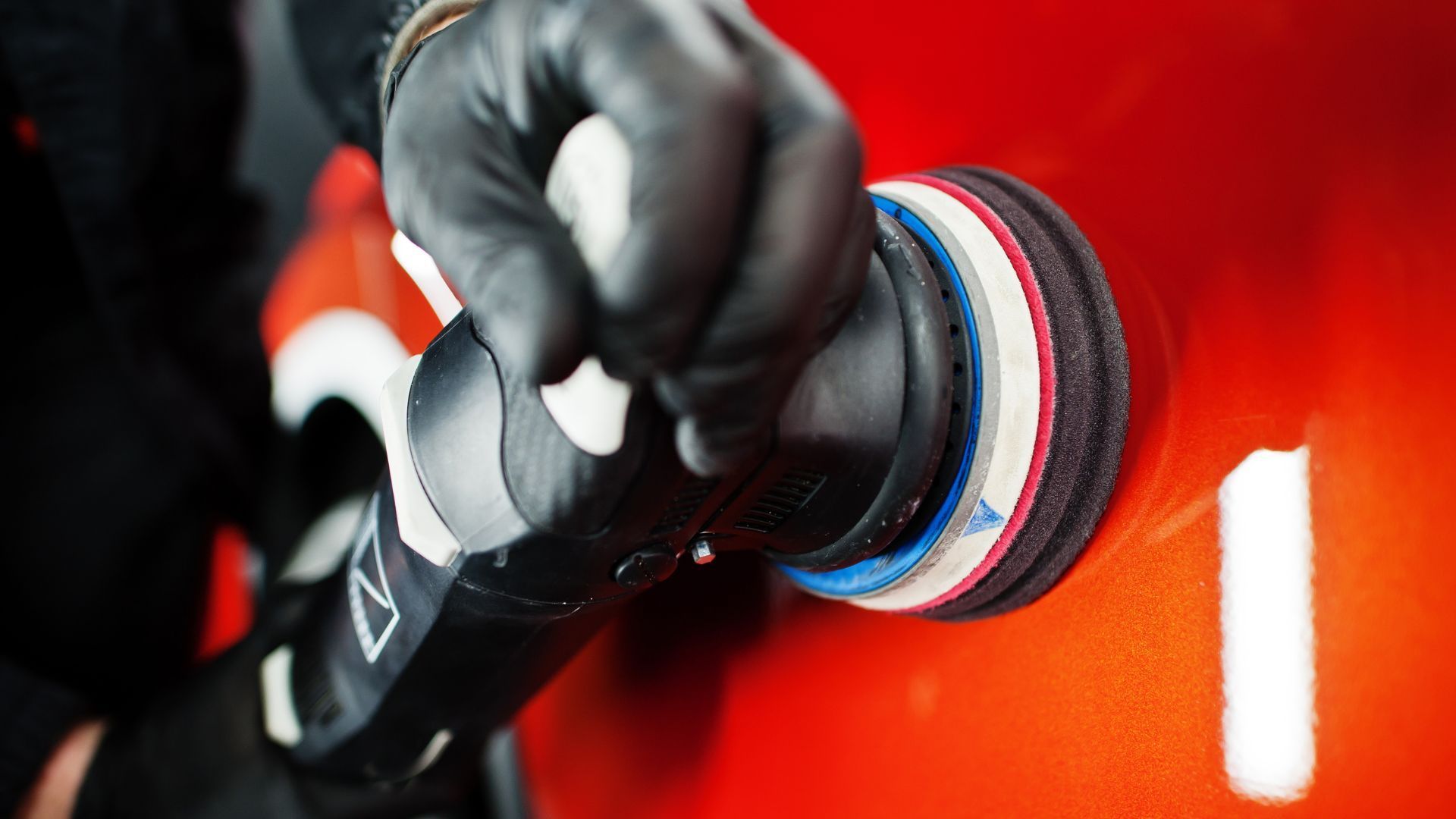 Close-up photo of a professional detailer in Duluth, GA, polishing a vibrant red car using a modern electric buffer. The detailer, wearing black gloves, expertly manipulates the buffer to achieve a high gloss finish. The focus is on the detailer's hands and the tool, emphasizing the shine and precision involved in the car detailing process, with reflections visible on the smooth, shiny surface of the car.