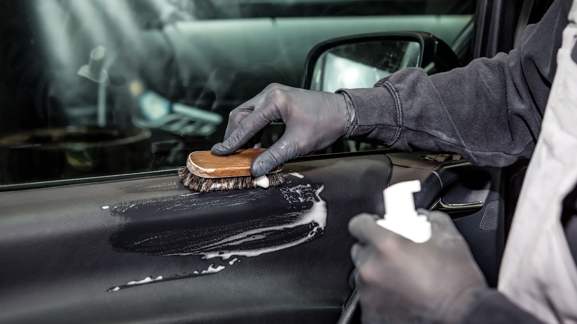 In Duluth, GA, a close-up shot shows a worker detailing a car's interior door. The worker, wearing black gloves and a dark sweatshirt, scrubs the door panel with a brush, creating lather with a cleaning solution. The door panel is black, and a vehicle's side mirror and window are visible in the background, slightly blurred. The worker holds a spray bottle in the foreground, suggesting a thorough cleaning process is underway, indicating attention to detail and care in vehicle maintenance.