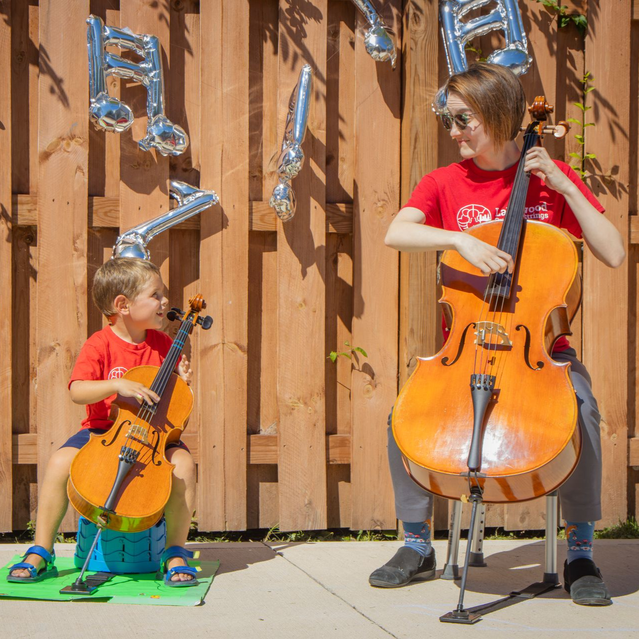 A young boy is playing a cello with his teacher in front of a fence